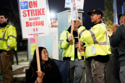 a young asian woman grips a sign that says on On Strike at Boeing. Others around her have signs. It’s nighttime.