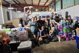 A group of fifteen workers pose inside a warehouse among boxes of diapers and other aid material