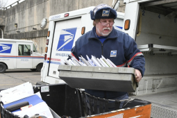 A man with a white beard in a blue postal uniform loads a tray of letters in to the back of a postal truck.