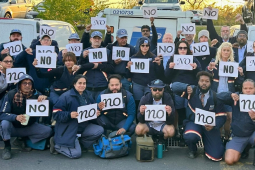 At least 32 letter carriers in uniform, holding "no" signs printed in various fonts, pose in a post office parking lot. The group looks diverse in race and gender. Several give the "thumbs-down" hand signal.