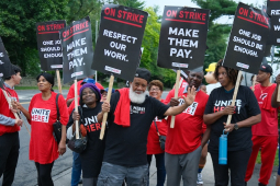 A group of a dozen hotel workers with signs saying “On Strike, Make them Pay” look at the camera, one waves