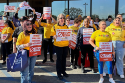 A crowd of people, mostly women, racially varied, in yellow T-shirts (some say UFCW, some say LIBRE) stand outside a grocery store, holding printed red-and-white signs, shouting together. There is a podium. Printed signs have UFCW Local 324 logo and such messages as "Stop the merger," "Say no to higher prices, fewer choices, store closures," and "Protect workers, protect customers, protect community." 