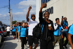 Workers, some in blue vests, march outside along an Amazon building. Some are holding their phones aloft, recording. Many are Black. They look like they are striding confidently and chanting. 