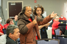 A person holding a mic stands speaking, gesturing with one arm, while others in the meeting listen intently. The group is diverse in race, gender, and age.