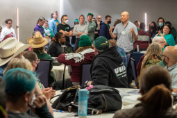 In a hotel conference room, one person stands and talks, while many seated at circular tables or standing against a wall listen thoughfully.