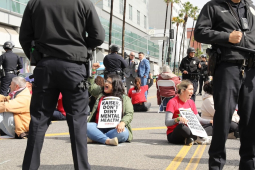 Several workers sit in the street with signs saying “Kaiser Don’t Deny Mental Health”--they are surrounded by police.