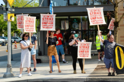 Eight picketers, who appear mostly young and white, stand in front of New Seasons. Their screen-printed picket signs say "New Seasons Labor Union, On Strike, Unfair Employer, Do Not Patronize" in red ink with flourishes. Two hold a black banner with yellow letters, only partly visible but appears to say "On strike." They all stand near the curb, facing traffic, and one has a bullhorn.