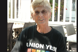 A smiling woman is pictured with short blond hair and a black Union Yes t-shirt with a rainbow on it.