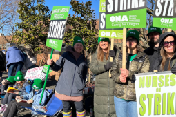 Women stand on a sunny day, wearing big jackets, holding green printed ONA picket signs that say "Unfair Labor Practice Strike" and "Nurses on Strike." A handmade sign visible in the background says "Burnout caused this turnout."