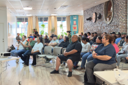 In a bright sunny room, a man in a black polo shirt and jeans stands on the left, speaking into mic, while about 30 people seated in chairs on the right listen. Most or all appear Latino/a. 