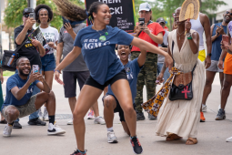 A Black dancer dances joyfully on the picket line, her long hair streaming out behind her. She is wearing a blue AGMA shirt. A delighted crowd, also mostly Black, surrounds her watching and taking photos -- some also wear AGMA shirts. The photo is full of energy, motion, and joy.