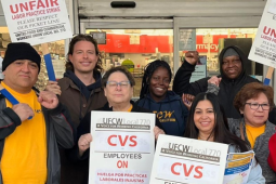 Eight people, varied in race and gender, pose together in front of a CVS store. Some wear UFCW yellow shirts and most carry picket signs that say "UFCW Local 770: CVS employees on unfair labor practices strike, please respect the picket line" in English and Spanish.