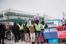 Workers, most of them Black, bundled against the cold, stand in front of an Amazon delivery facility sign. Their banner says, in French, "Without us, nothing is free" and the name of the union. Many hold CSN flags, and smile resolutely. On the right, a figure viewed from back silhouette speaks to them, one hand pointing the way forward.