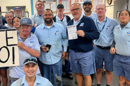 Seventeen uniformed letter carriers pose together in a post office, holding a big handmade "VOTE NO!" sign.