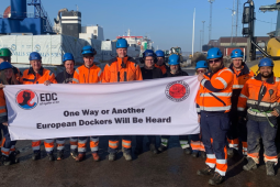 Fourteen workers in orange vests, blue hardhats, and workboots stand in a port in bright sunlight. They hold a big white banner that says "One way or another, European dockers will be heard" with logos of the SDU and the European Dockworkers Council. Most but not all appear to be white men.