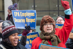 A crowd of federal workers stand outside, bundled against cold, carrying printed AFGE signs that say "EPA employees deserve respect" and "Stop the shutdown." In the foreground are a Black woman and a white woman, the latter raising a red-gloved fist in the air, both chanting or singing.