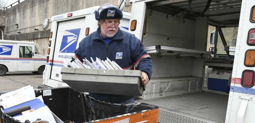 A man with a white beard in a blue postal uniform loads a tray of letters in to the back of a postal truck.