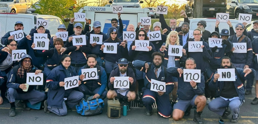 At least 32 letter carriers in uniform, holding "no" signs printed in various fonts, pose in a post office parking lot. The group looks diverse in race and gender. Several give the "thumbs-down" hand signal.