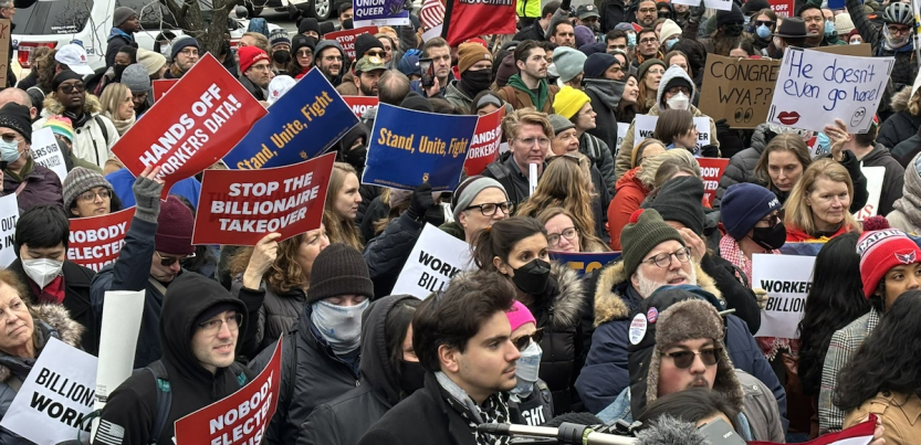 A sea of faces in a protest crowd. Some printed signs say "Stop the Billionaire Takeover," "Hands Off Workers Data," "Stand, Unite, Fight," and "Nobody Elected Musk." Some handlettered signs say "Congress, WYA?" and "He doesn't even go here!"