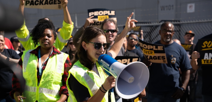 Several workers in green high-viz vests chant and hold signs, one woman in front speaks into a megaphone.