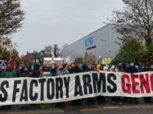  A large crowd of people bundled against the cold, some masked, hold a long banner: "THIS FACTORY ARMS GENOCIDE." A few small handmade signs are also visible: "Ceasefire now, end the occupation," "Jews against genocide," and a Palestinian flag. Behind them is the factory with a big "Eaton" logo.  