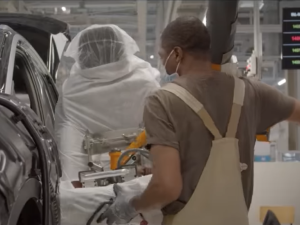 A masked worker in overalls place a seat into a car on an assembly line.