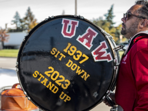 A UAW member walks with a drum that reads "UAW: 1937 - Sit-Down, 2023 - Stand Up."