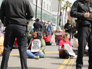 Several workers sit in the street with signs saying “Kaiser Don’t Deny Mental Health”--they are surrounded by police.