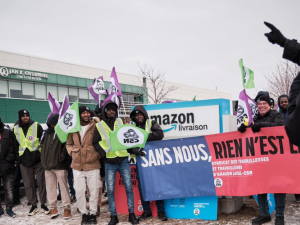 Workers, most of them Black, bundled against the cold, stand in front of an Amazon delivery facility sign. Their banner says, in French, "Without us, nothing is free" and the name of the union. Many hold CSN flags, and smile resolutely. On the right, a figure viewed from back silhouette speaks to them, one hand pointing the way forward.