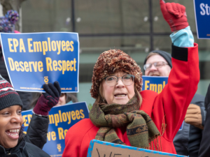 A crowd of federal workers stand outside, bundled against cold, carrying printed AFGE signs that say "EPA employees deserve respect" and "Stop the shutdown." In the foreground are a Black woman and a white woman, the latter raising a red-gloved fist in the air, both chanting or singing.