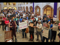 A group of 80 people with signs stand inside a collegiate-looking building. A woman in front holds a sign saying “Bioengineering for a Fair Contract.”