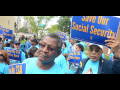 A crowd dressed in light blue T-shirts holds signs saying Save our Social Security and Keep My SSA Office Open.