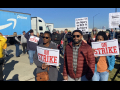 A group of workers and supporters hold signs that read STRIKE in front of a blue Amazon Prime truck.