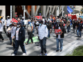 Masked people marching outside. Some have signs: "UNLOCK THE MONEY," "FREE OUR FUNDING," "AUSTERITY IS RACIST!"