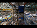 An aircraft factory floor is viewed from above, with planes on either side and high rafters above.