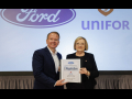 A man and a woman in suits stand holding a binder together in front of a screen that has Ford and Unifor logos projected on it.