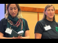 Two women in dark t-shirts stand side by side, one reading from a thick booklet