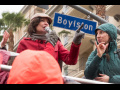 A woman speaks at a strike rally while another woman interprets in sign language. Behind them is a palm tree, an apartment buidling or hotel, and a street sign "Boylston"