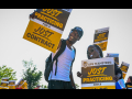 A young slender Black man in a baseball cap smiles down at the camera with a sign saying “Just Practicing for a Just Contract.” To the left of him is a Black woman in sunglasses with a whistle in her mouth carrying the same sign..