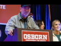 A man in a ball cap stands behind a lectern that says Osborn for Senate