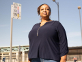 Black woman looks into the distance with a serious, determined expression. Behind her is a bus stop sign and a blue sky.