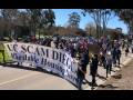 A group marches down the street behind a banner reading "UC Scam Diego: Affordable Housing Now."