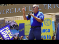 Man in blue shirt with right arm raised and left arm holding microphone addressing crowd, Fuera LUMA flag visible in foreground, building of Puerto Rico Electric Power Authority (PREPA)