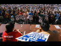 In the foreground we see the backs of a few strikers in red UAW 2865 T-shirts, one holding a strike sign, sitting at a table. Facing them is a huge crowd of strikers, standing, with picket signs, outdoors. The signs say "UAW on strike, unfair labor practice."