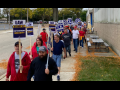 A dense line of picketers (at least 25 people are visible but it looks like there are more behind them) marches towards the camera on a leafy sidewalk alongside a factory. Many carry printed signs like "UAW on strike" and "Stand up for retirement security." Most visible faces are white but a few Black faces are also visible; there's a mix of women and men.