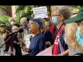 A group of older people in masks stands outside. The person in the middle is speaking into a mic. Someone behind her holds up a printed sign: "City Retirees Say NO, Don't Privatize Our Senior Care!"