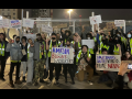 Crowd of excited owrkers stands outside warehouse in the dark holding up handmade signs. Most prominent ones read "Amazon burns workers," "Amazon exploits workers," "Amazonians unite! Without us they're nothing."