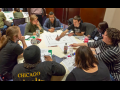 People sit around a big table, leaning in. One is drawing a workplace map on big paper, while another points something out. They're in a hotel conference room. Other table groups are visible behind them.