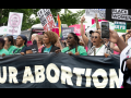 Women march behind a giant banner. The part we can see reads "OUR ABORTION." Other signs visible say "If it's not your body, it's not your choice" and "Our bodies, our future."