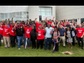 A large group of Black and white women and men stands on grass outside the Mercedes-Benz Training Center, smiling, many with fists in the air. Many wear red T-shirts with a UAW logo and the words "Mercedes Workers United."